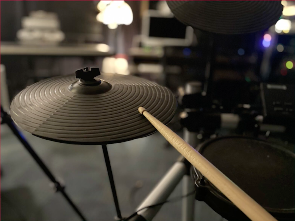 A drumstick strikes an electronic cymbal pad on a digital drum set, with blurred studio equipment visible in the background.