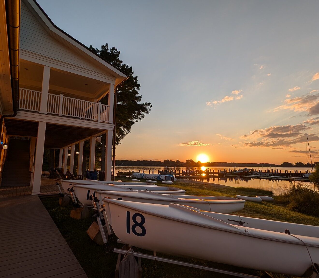 A row of small sailboats on trailers next to a building overlooking a lake at sunset.