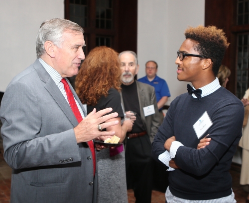 Two men standing and conversing at a formal indoor event, while other attendees mingle in the background.