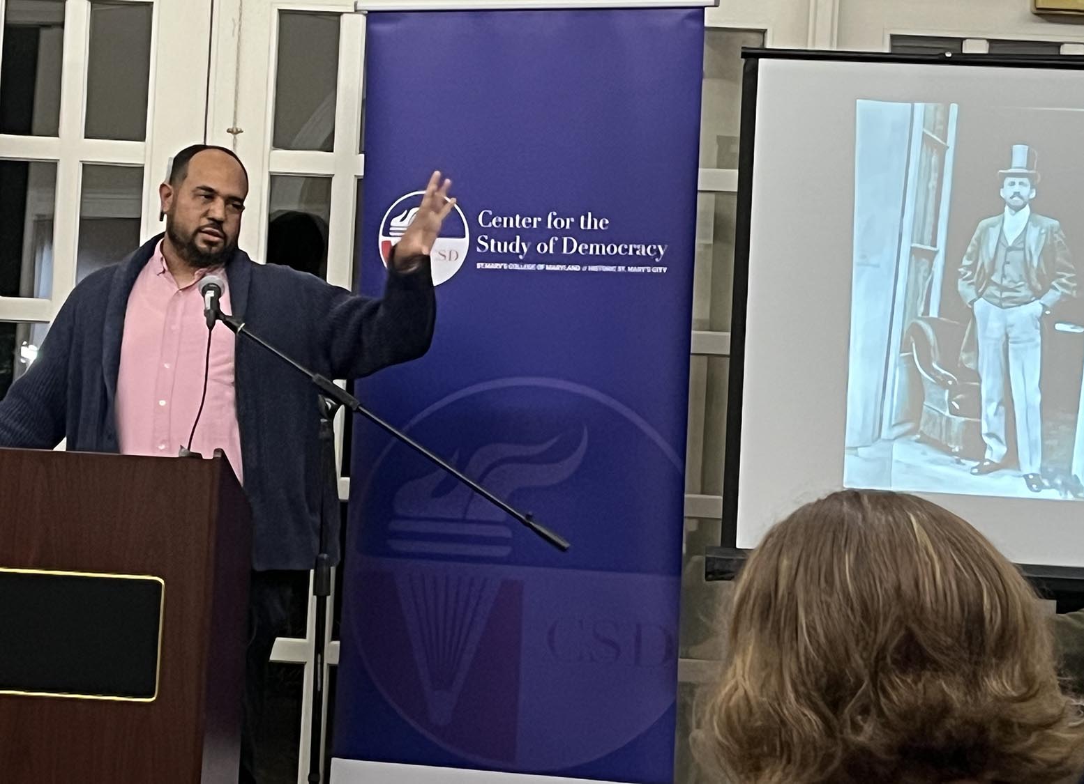Aaron Yates speaks at a podium with a microphone, in front of a Center for the Study of Democracy banner. A projector screen shows a vintage black-and-white image of a man in historic attire, with the audience visible from behind