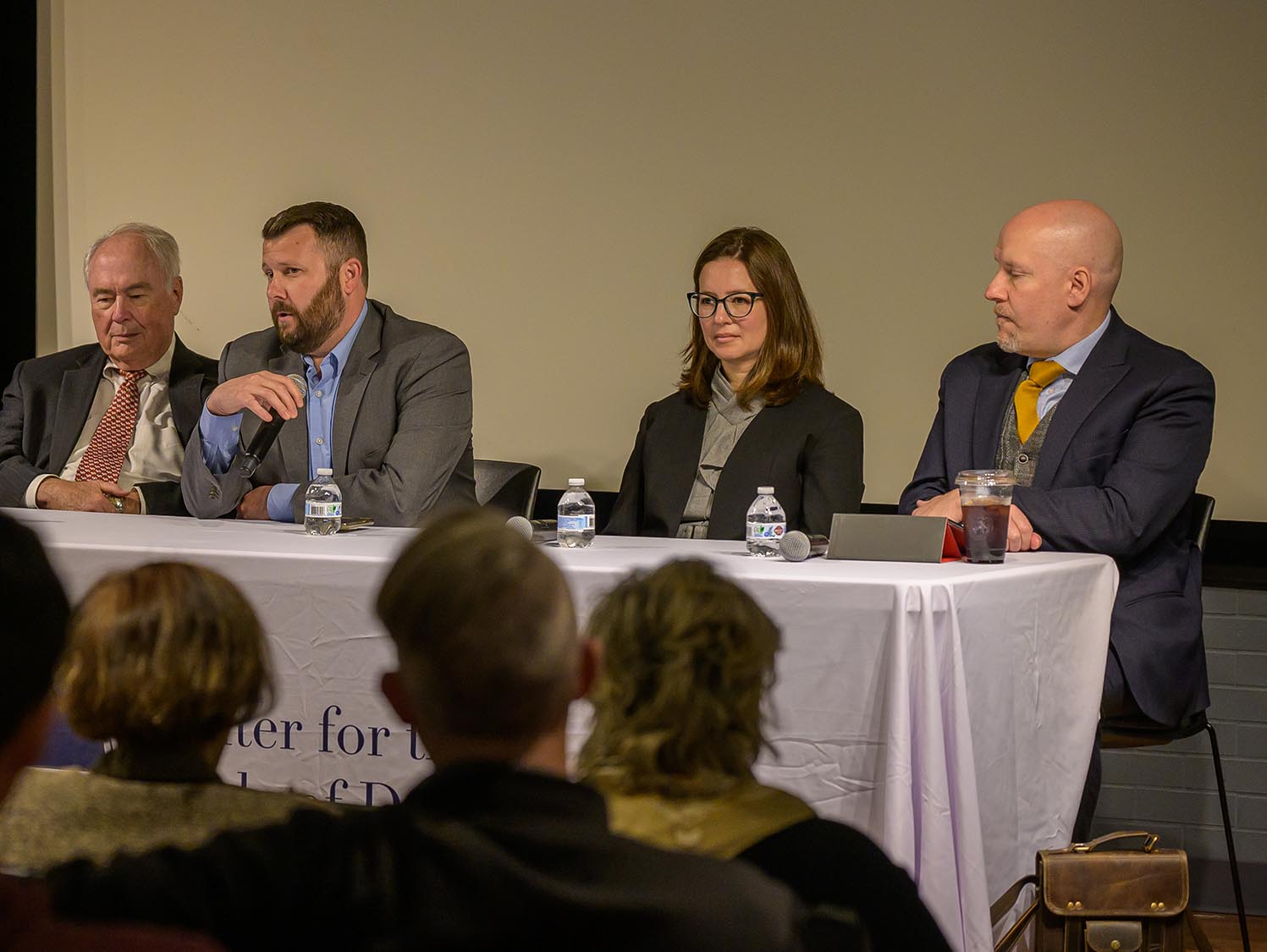 Four people are seated at a table on a stage, participating in a panel discussion. Two men and a woman are speaking, while one man is quietly observing. A white tablecloth with text is partially visible. Audience members are in the foreground.