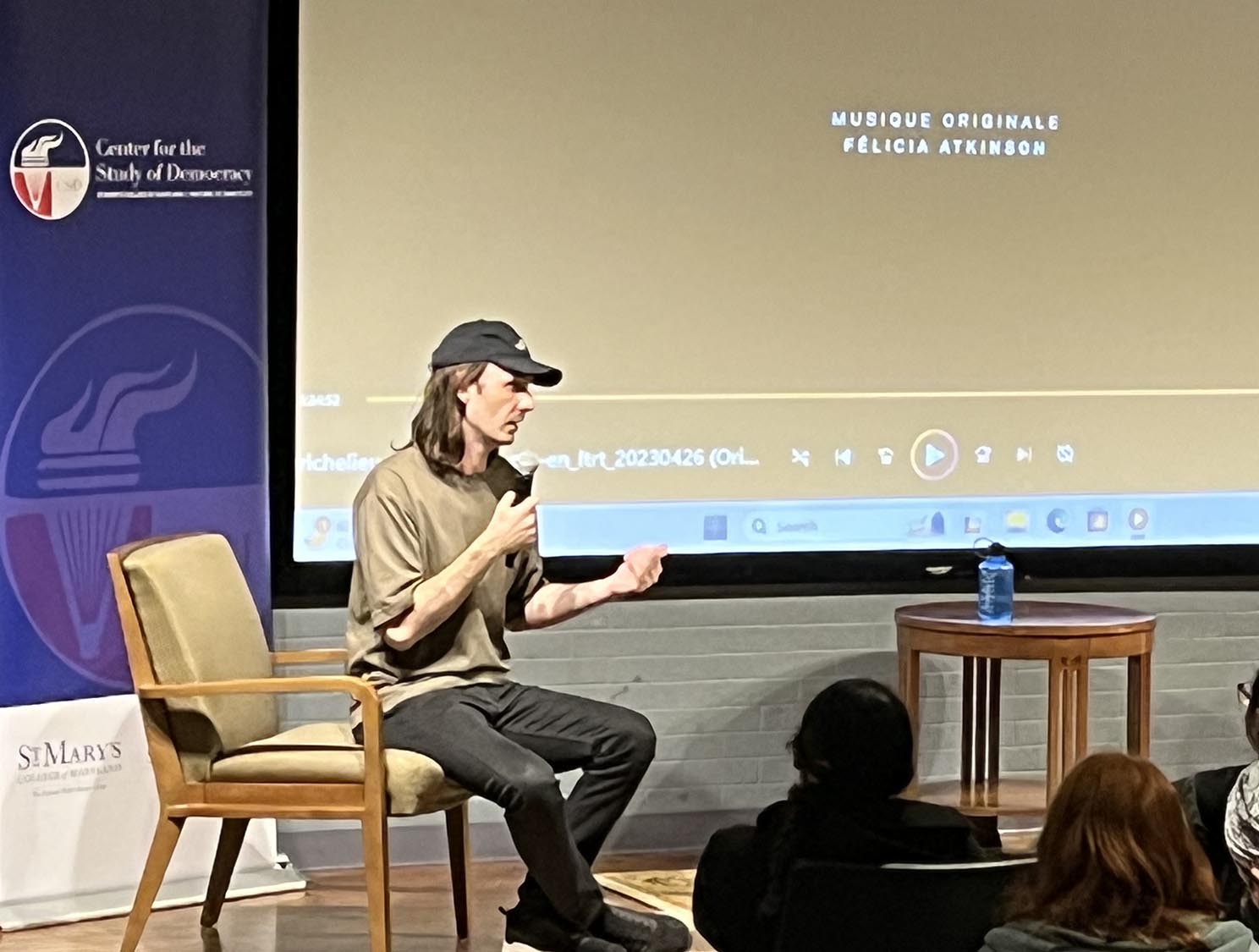  APier-Philippe Chevigny wearing a cap and holding a microphone sits on a chair, speaking to an audience.