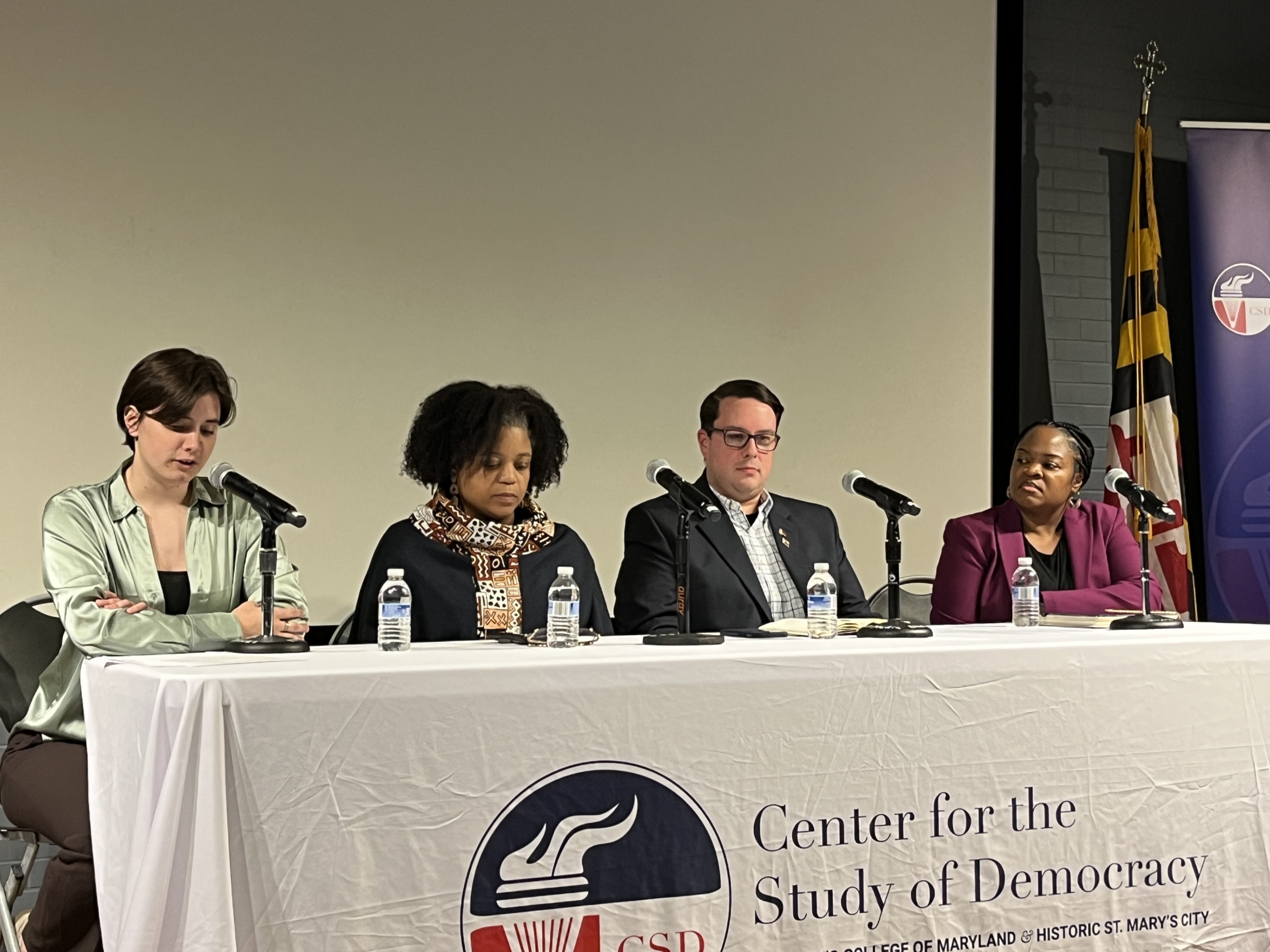 Four panelists sit at a table with microphones and water bottles during a Center for the Study of Democracy event. A Maryland state flag is visible in the background.