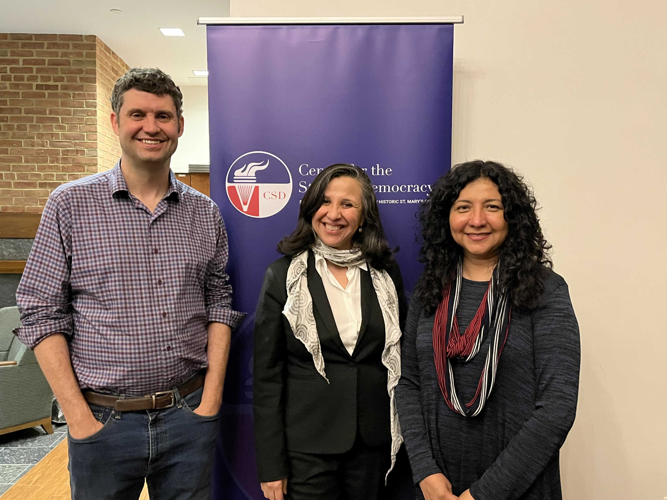  Three adults stand in front of a blue banner that reads “Center for the Study of Democracy.” Two women and one man are smiling at the camera.