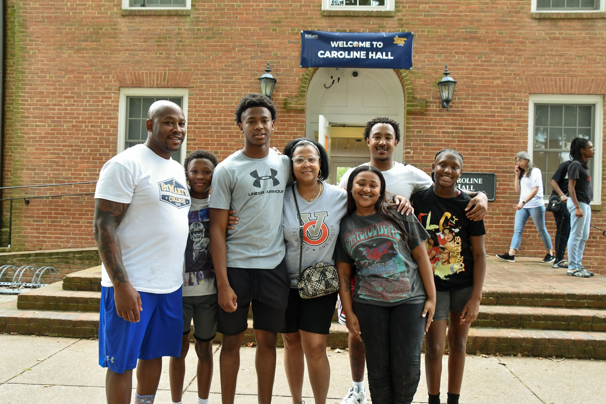 A group of seven people pose and smile in front of Caroline Hall, a brick building with a “Welcome to Caroline Hall” sign above the entrance.