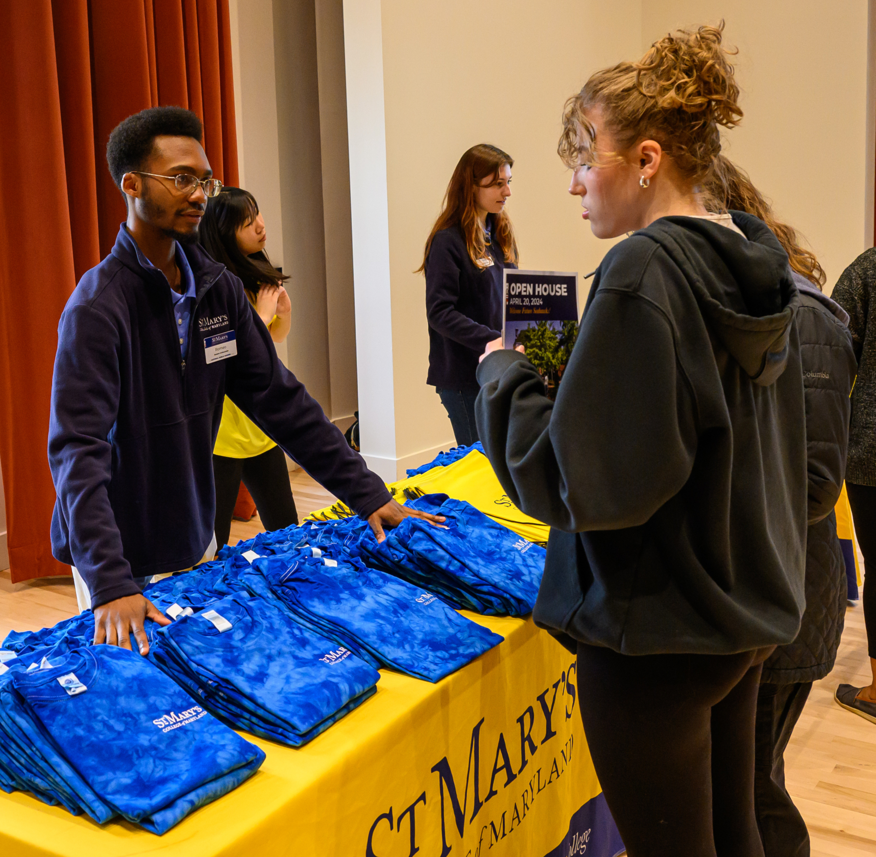 A man stands behind a table with blue drawstring bags, speaking to a woman at a St. Mary’s College of Maryland event. Other people are visible in the background.