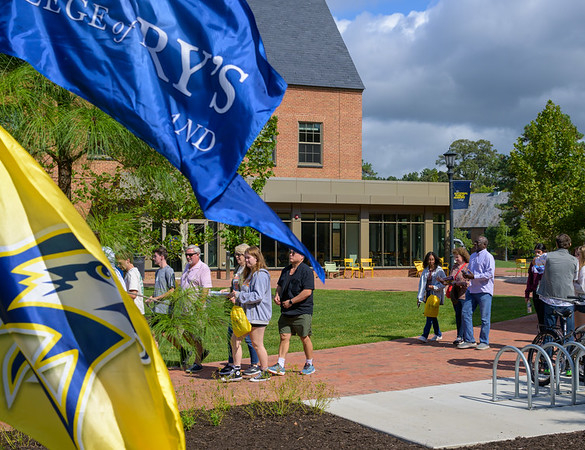 54013208639_89f4a4bf61_c People walk and gather outside a college building on a sunny day, with blue and yellow flags in the foreground.