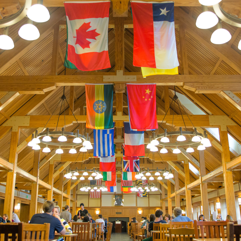 A vibrant dining hall with wooden beams and high ceilings, decorated with various international flags, where students sit together and enjoy meals at wooden tables.