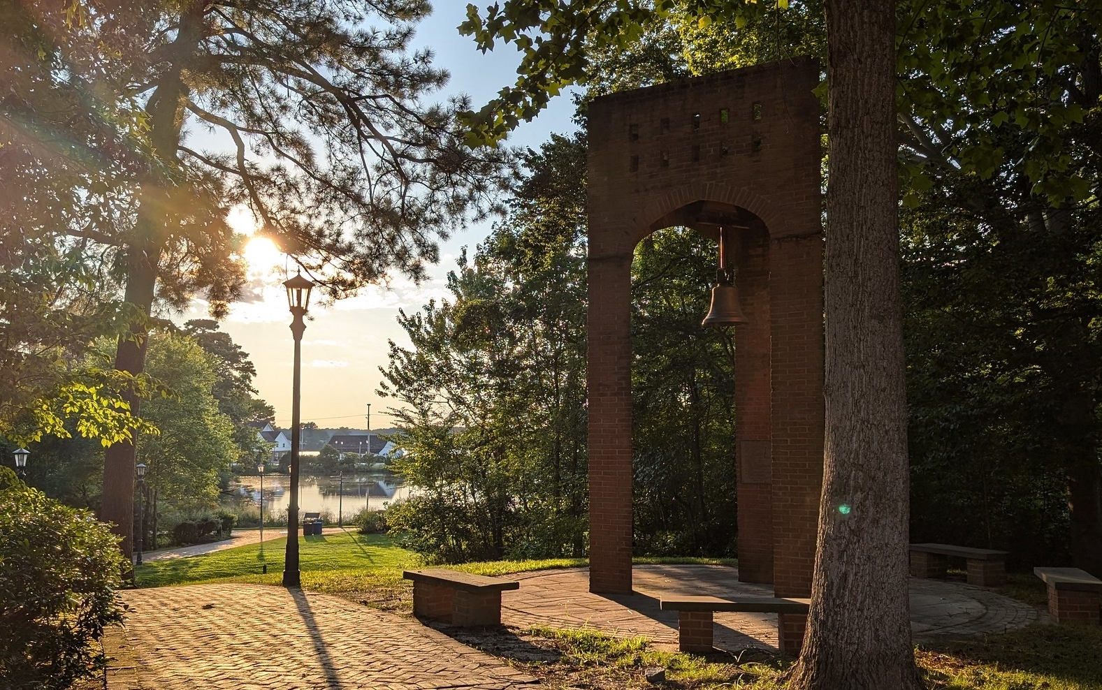 Brick arch structure with a bell, surrounded by trees and benches, sits near a paved walkway at sunset with a river visible in the background.