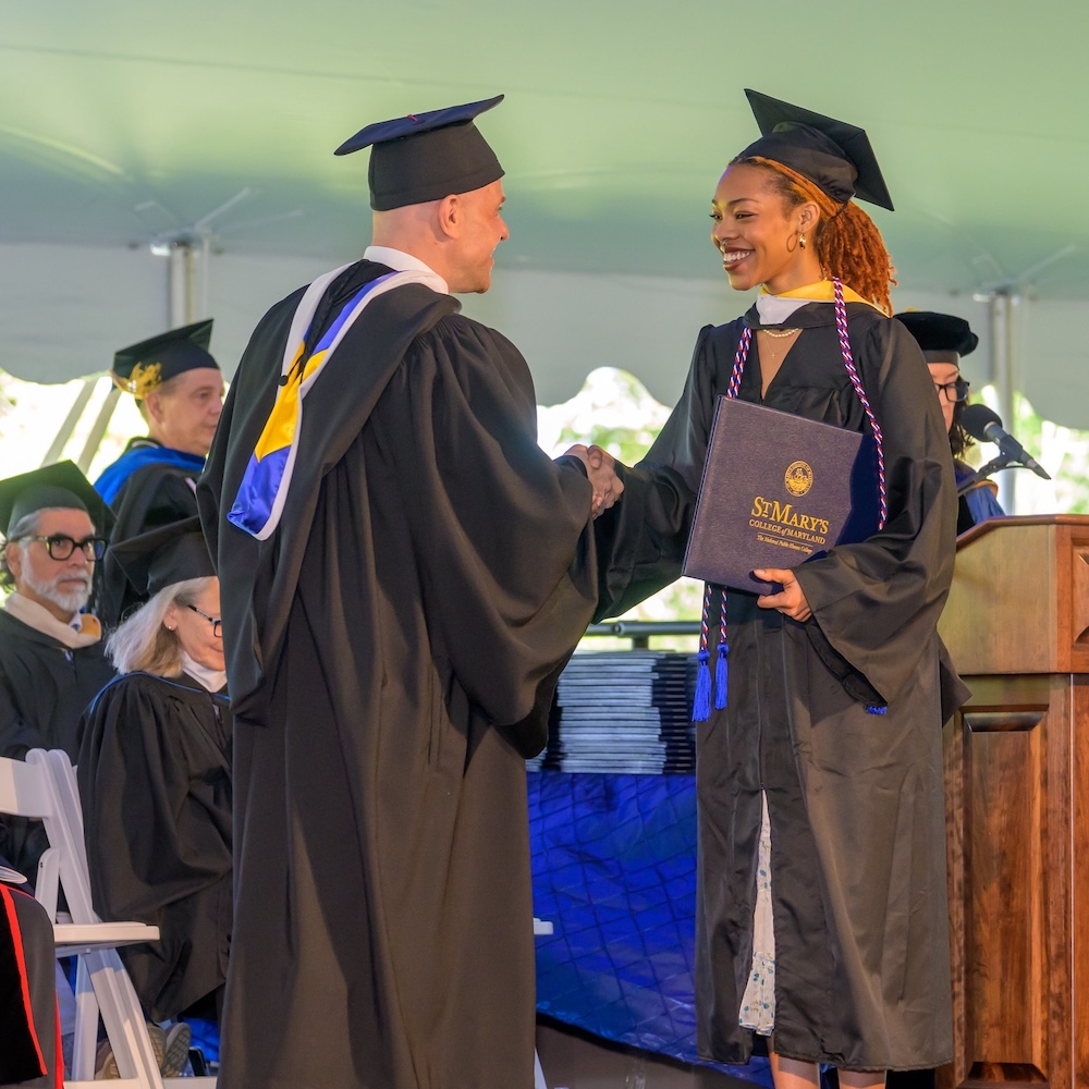 A student in a cap and gown shakes hands with a college administrator while receiving a diploma at a graduation ceremony.