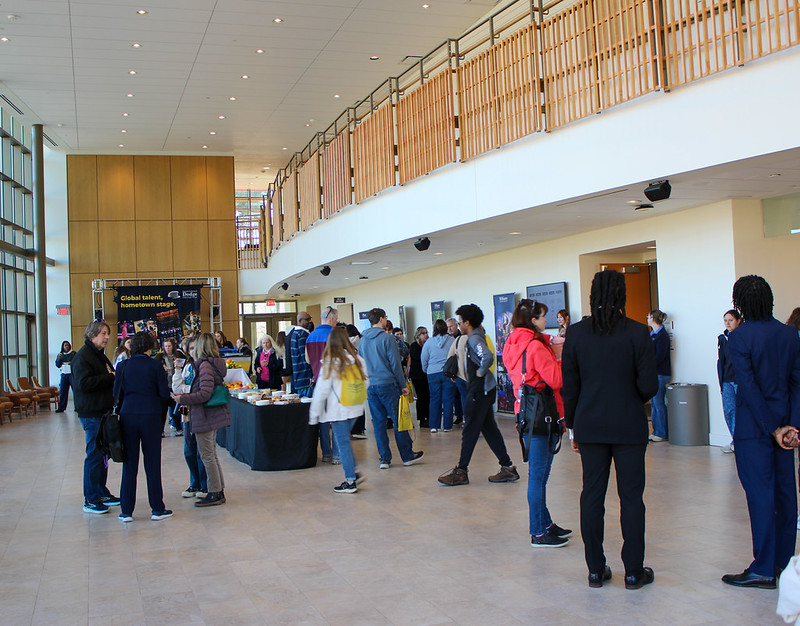 People gather and socialize in a spacious, modern indoor atrium with tables of refreshments and informational displays in the background.