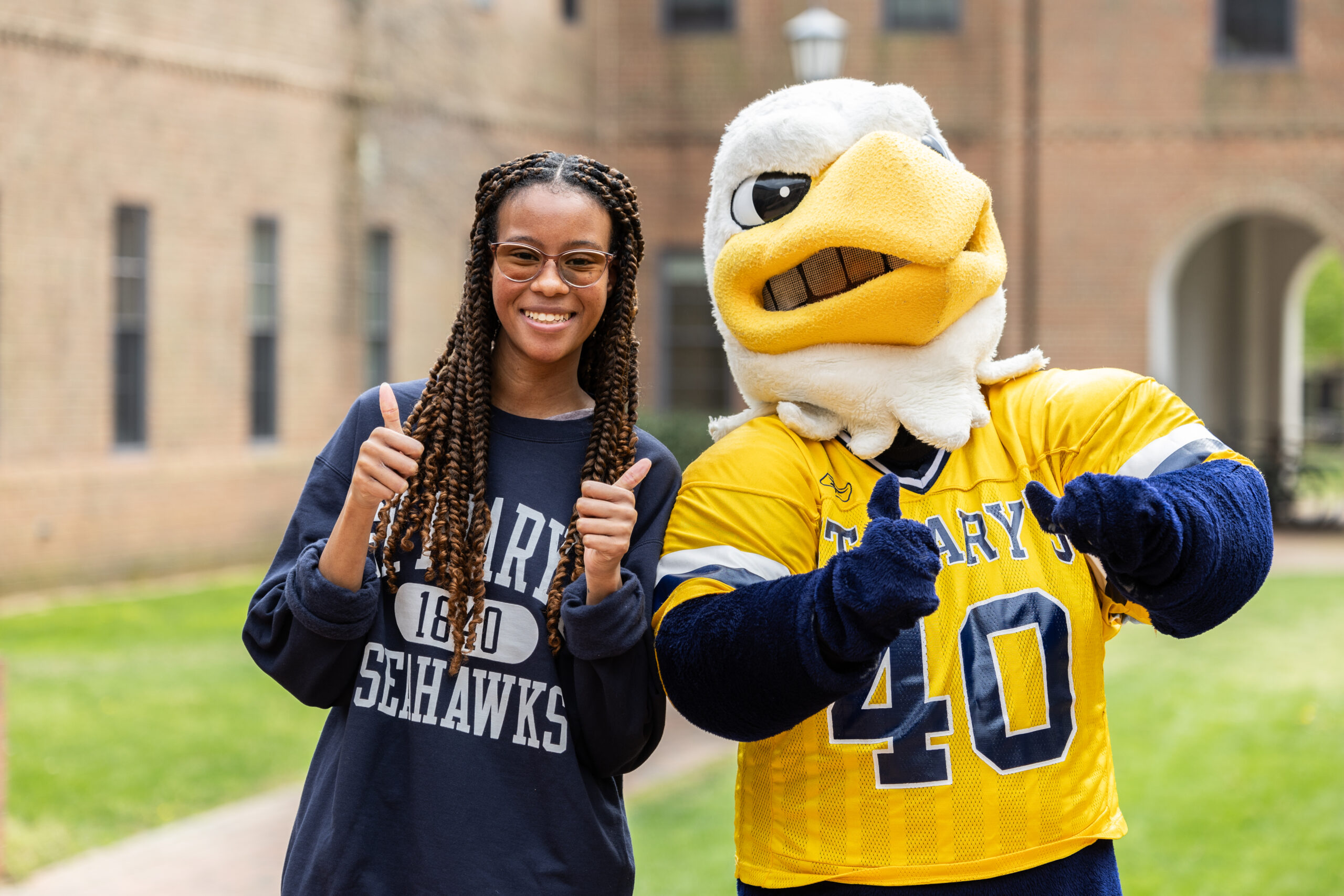 A female student in a navy sweatshirt and a school mascot in a yellow jersey both giving a thumbs up.