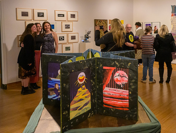 People view artwork and socialize at an indoor gallery exhibition featuring paintings, framed photos, and textile art on the walls.