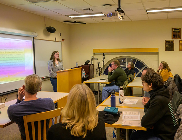 A person stands at a podium presenting to a group of people seated at tables in a classroom with a projection screen and papers in front of attendees.