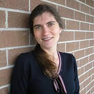 Alla Semenova A person with long brown hair and a black top, standing against a brick wall adorned with awards, smiling at the camera.