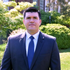 Antonio Ugues portrait A person in a dark suit and blue tie stands outdoors, an aura of prestige surrounding them, as if ready to accept awards against the backdrop of lush greenery.