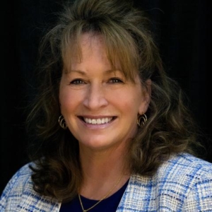 Cynthia Greb portrait Smiling woman with wavy hair and an award-winning style dons a blue and white plaid jacket against a dark background.