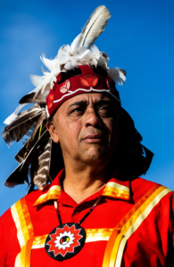 Francis Gray portrait A person in a red shirt and feathered headdress stands proudly against the blue sky.
