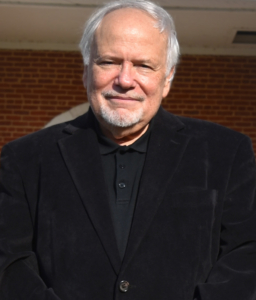 Jeff Silberschlag1 A distinguished man with white hair and a beard, dressed in a black jacket and shirt, stands proudly in front of a red brick wall, reminiscent of an awards ceremony backdrop.