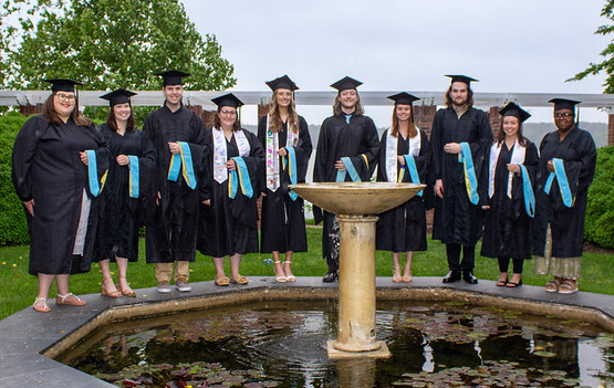 Ten graduates in caps and gowns stand in a line behind a fountain, each holding a blue and yellow sash, with greenery in the background.