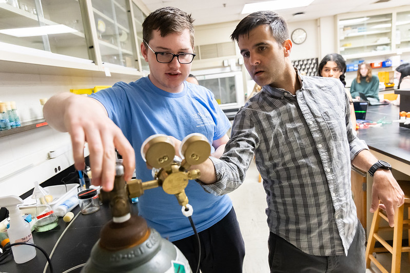 Two people adjust a gas cylinder regulator in a laboratory setting.