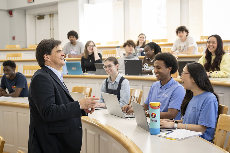 A professor stands and smiles while speaking to a group of students seated in a tiered classroom, some with laptops open and all appearing engaged.