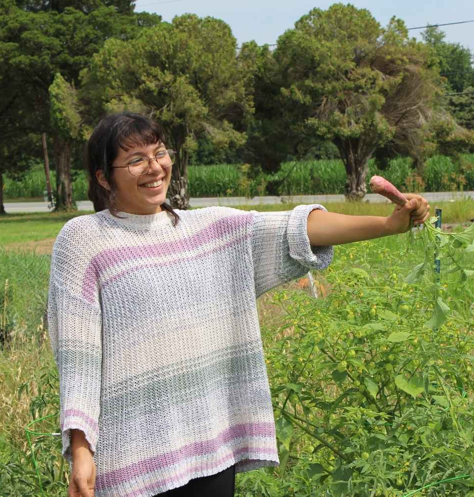 53068054174_daab5767e5_o A person standing in a green field, smiling and holding up a freshly harvested purple radish. Trees and a road are visible in the background.