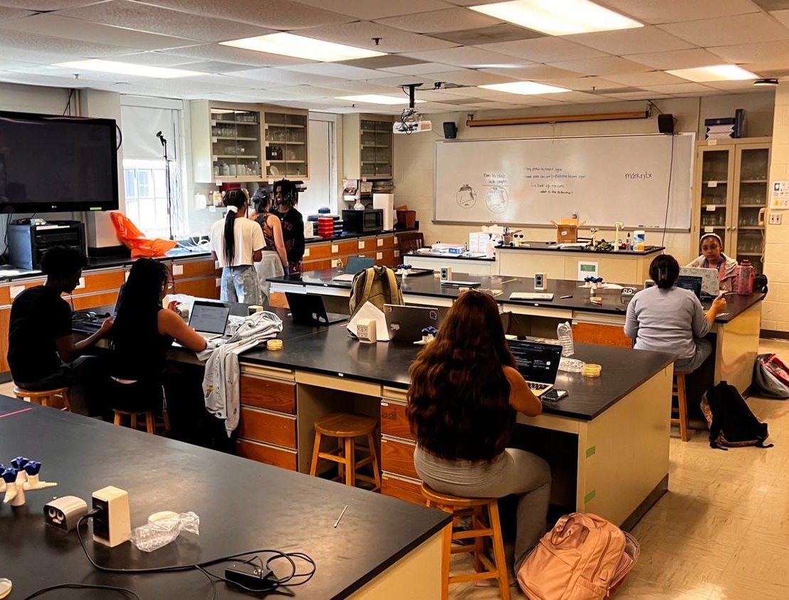 IMG_8537 Students sit at lab tables in a science classroom with equipment and laptops; a teacher stands at the front near a whiteboard and cabinets line the walls.