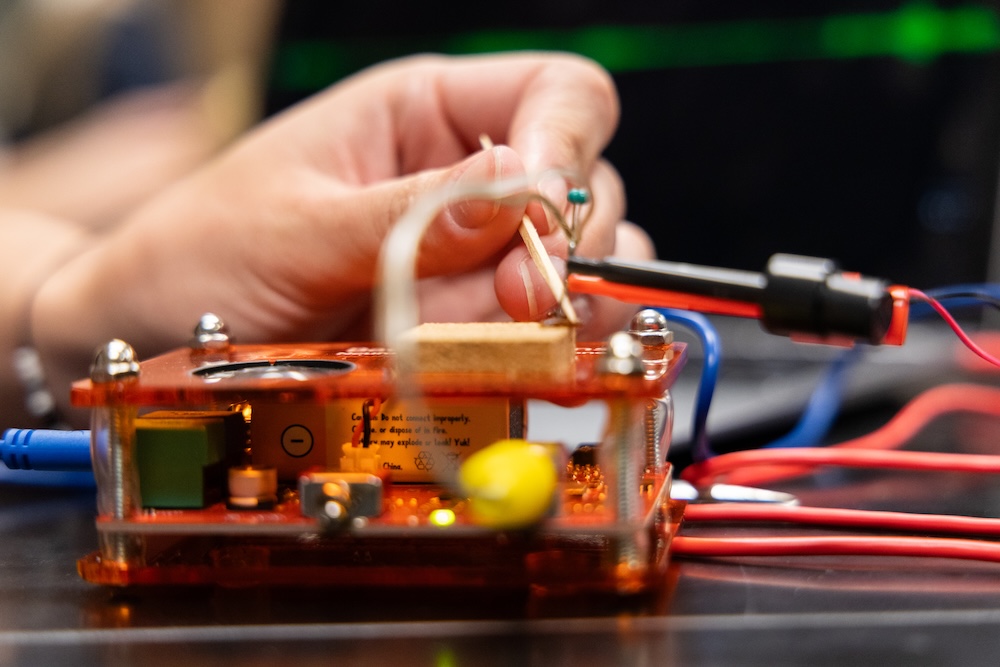 A hand adjusts wires on an electronic circuit board with various components and colored cables on a table.