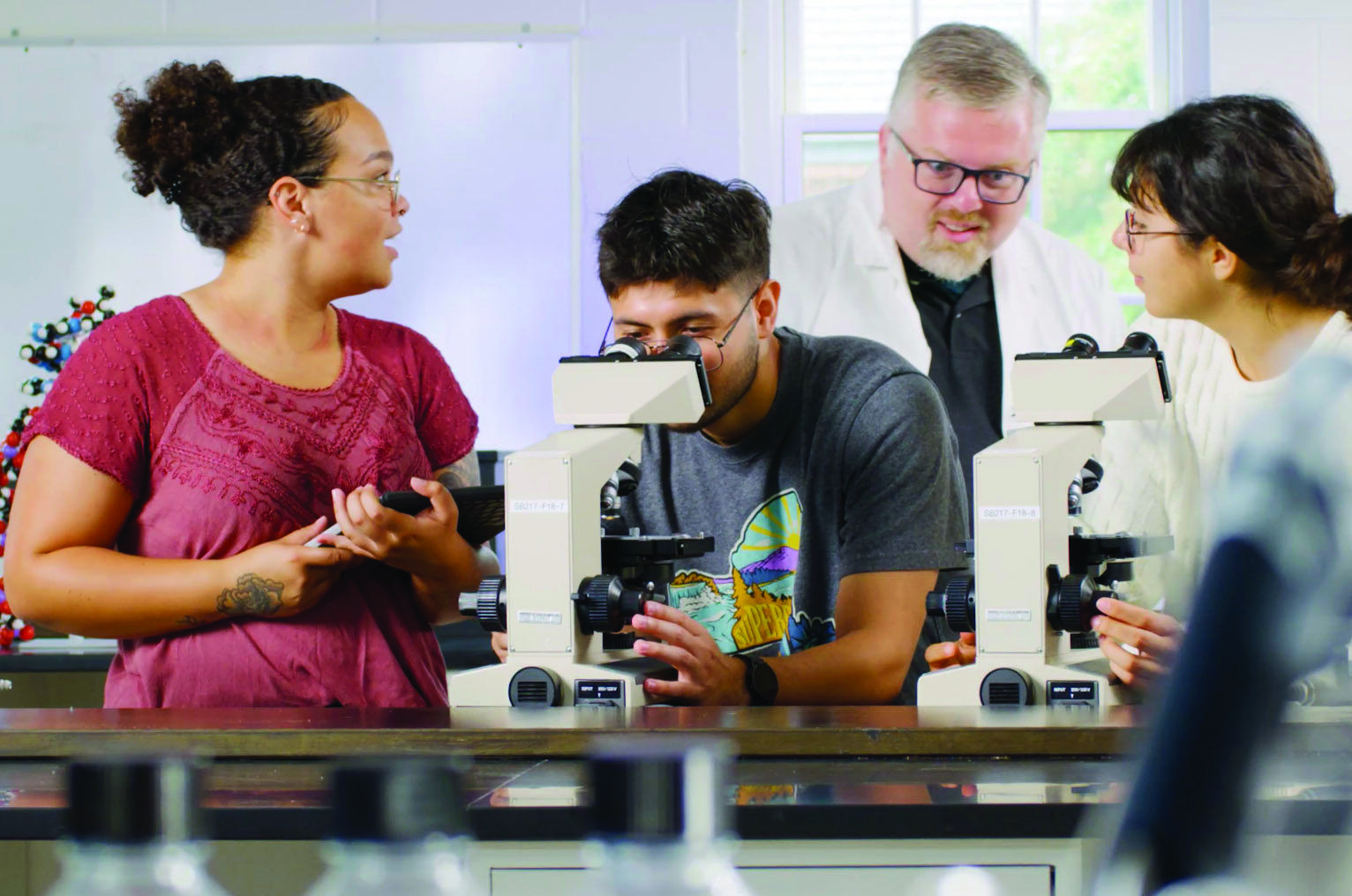Four people in a laboratory use microscopes and have a discussion; laboratory equipment and bottles are visible in the foreground.