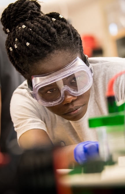 A person wearing safety goggles and gloves focuses intently while working with laboratory equipment in a science lab.