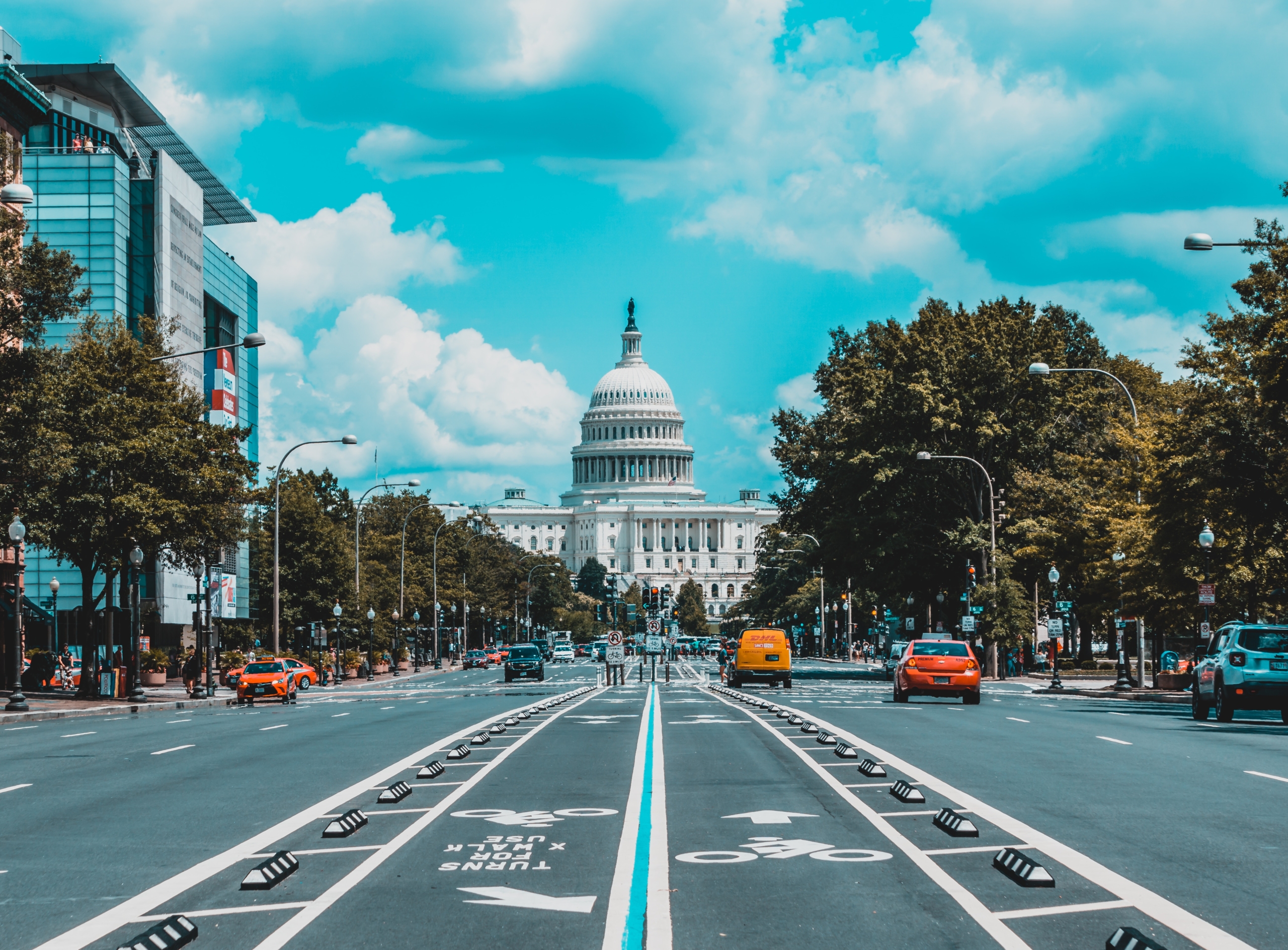 A view of the United States Capitol building from the middle of a divided street with bike lanes, trees, and cars under a partly cloudy sky.