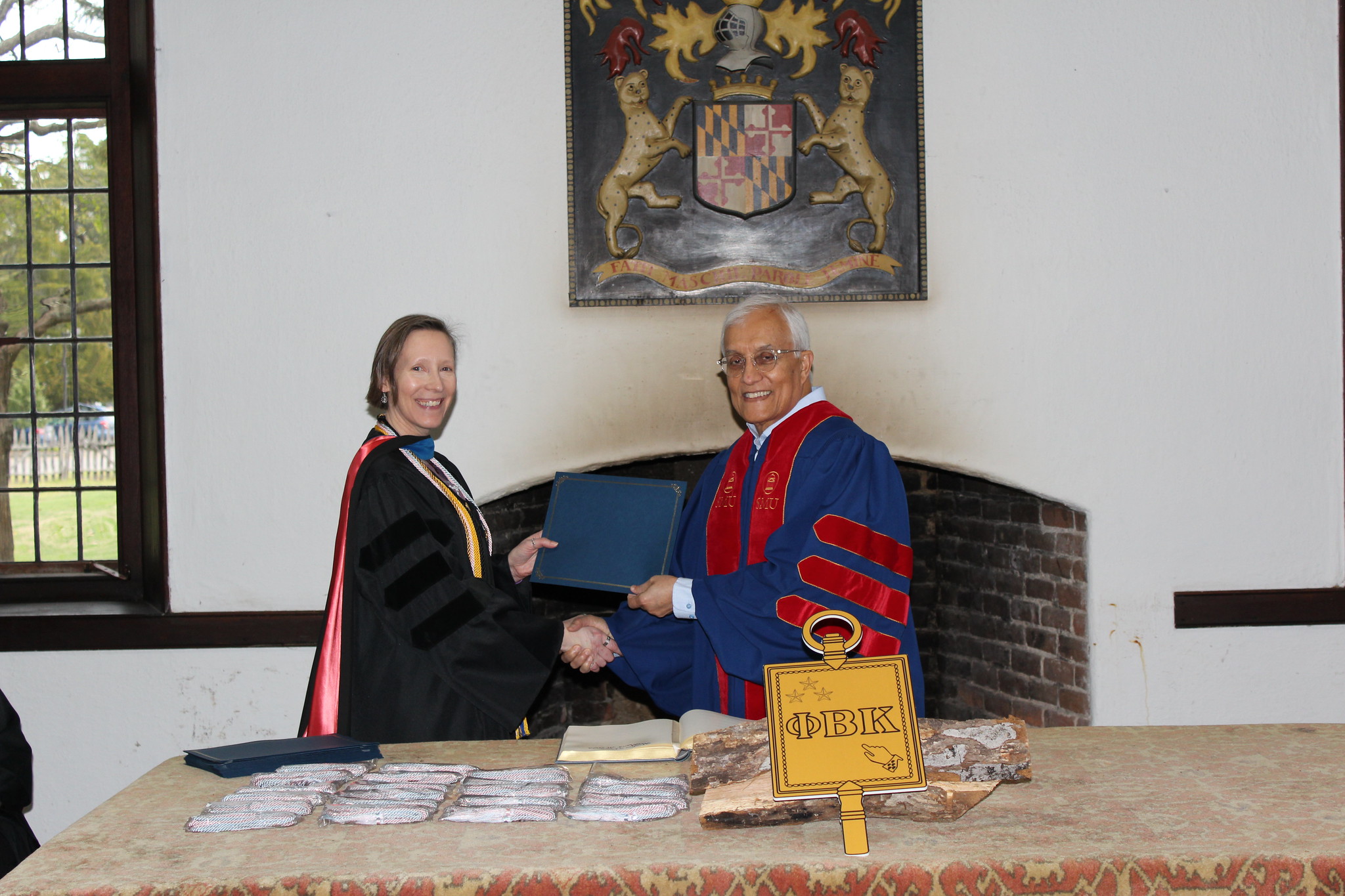Two people in academic robes shake hands and exchange a document in front of a fireplace, with a Phi Beta Kappa symbol and several stacked certificates on the table.