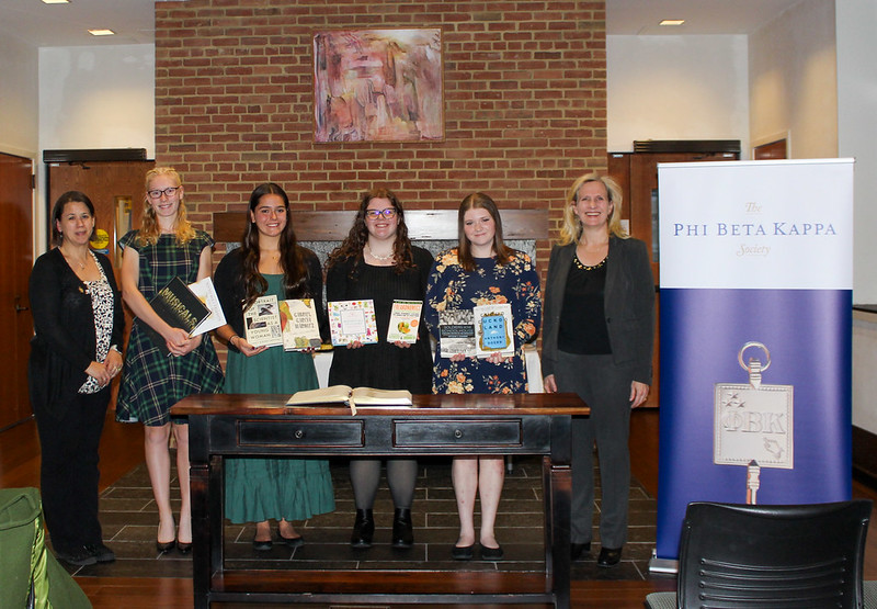 Seven women stand indoors behind a wooden desk; five in the center hold books. A Phi Beta Kappa Society banner is displayed on the right.