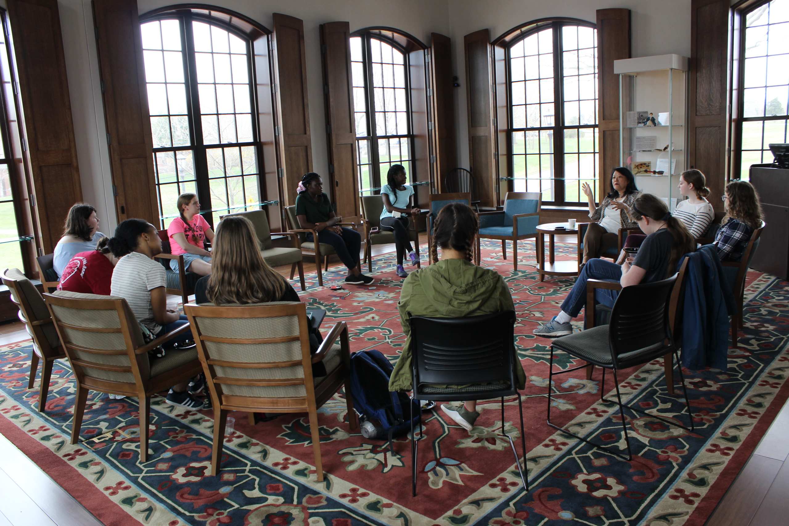 A group of people are seated in a circle on chairs in a well-lit room with large windows, having a discussion.