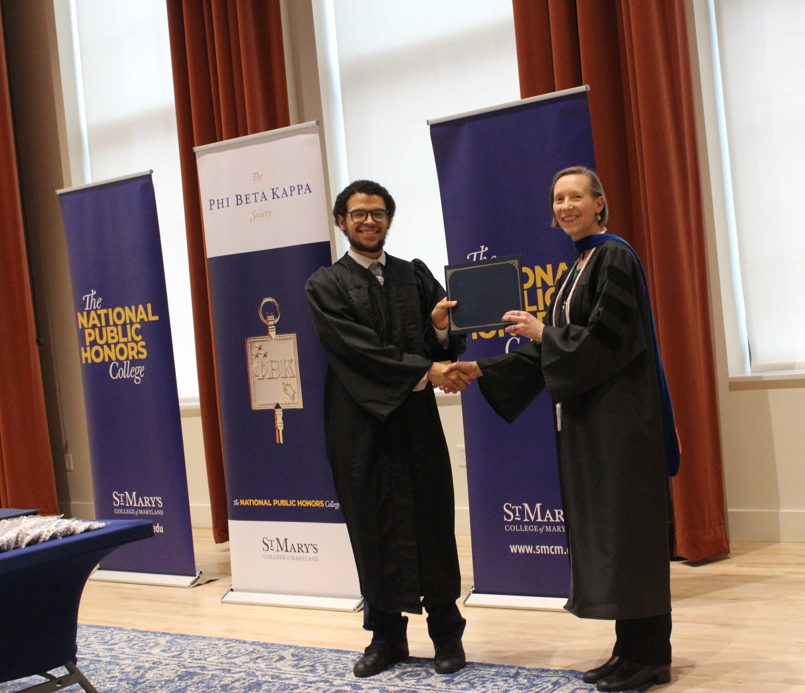 A student in a cap and gown receives a certificate from a faculty member at an indoor academic award ceremony.