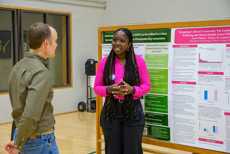 A woman presents research on a poster board to a man in a gymnasium setting. The posters display graphs, text, and scientific information.