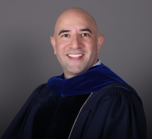 A man in academic regalia, featuring a dark blue velvet hood and gown, smiles at the camera against a plain gray background.