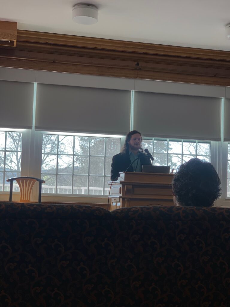 A person stands at a podium speaking in a room with large windows and closed blinds; two people sit in the audience facing the speaker.