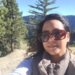A woman wearing sunglasses and a beige vest takes a selfie outdoors with pine trees and mountains in the background.