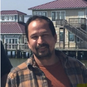 A man wearing a brown plaid shirt, proud of his recent awards, stands outdoors in front of waterfront houses on stilts under a clear sky.