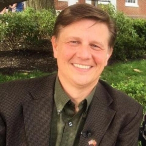 A man with short brown hair wearing a dark blazer and green shirt, smiles while sitting outdoors in front of bushes and a building.