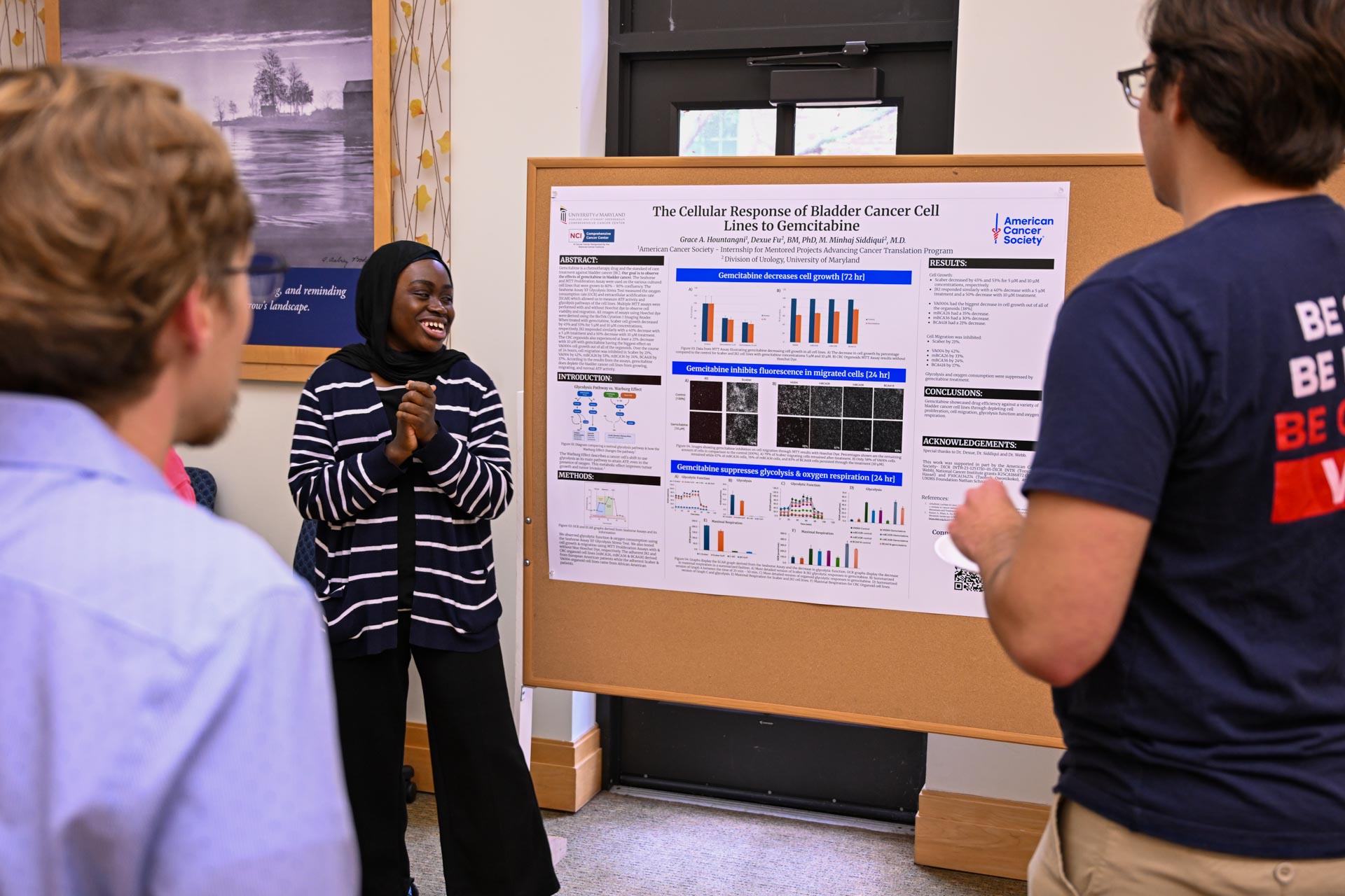 A woman presents research on bladder cancer cell lines shown on a large poster to three attentive people in a brightly lit room.