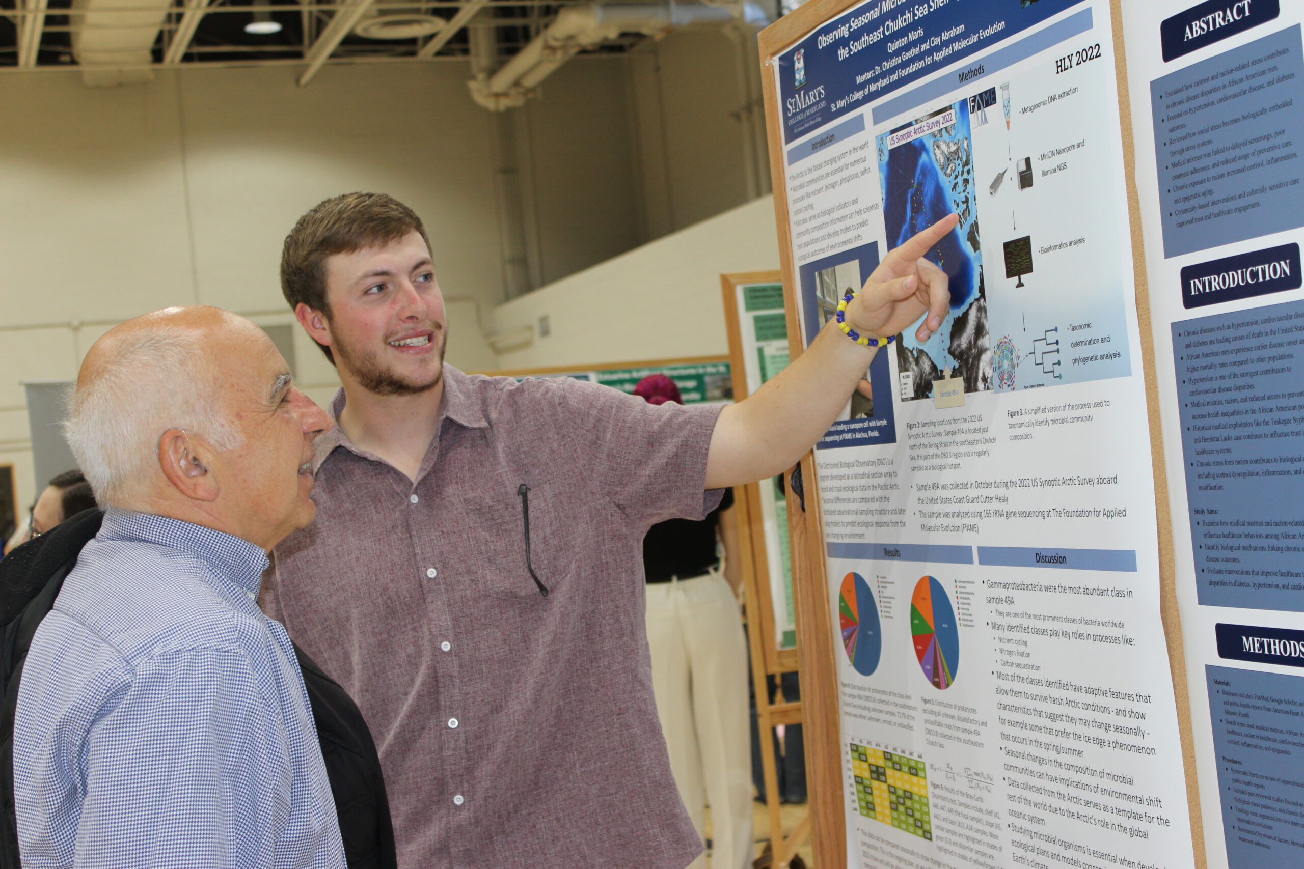 Two men stand in front of a scientific research poster; the younger man is pointing at the poster and explaining its content to the older man.