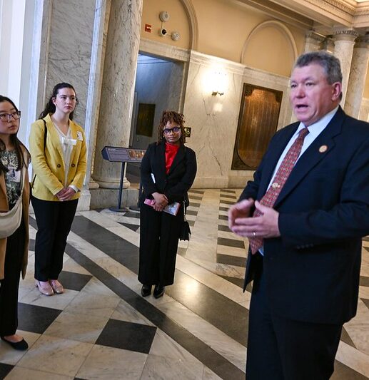 A man in a suit speaks to a group of people standing in a marble hallway with columns and ornate details.