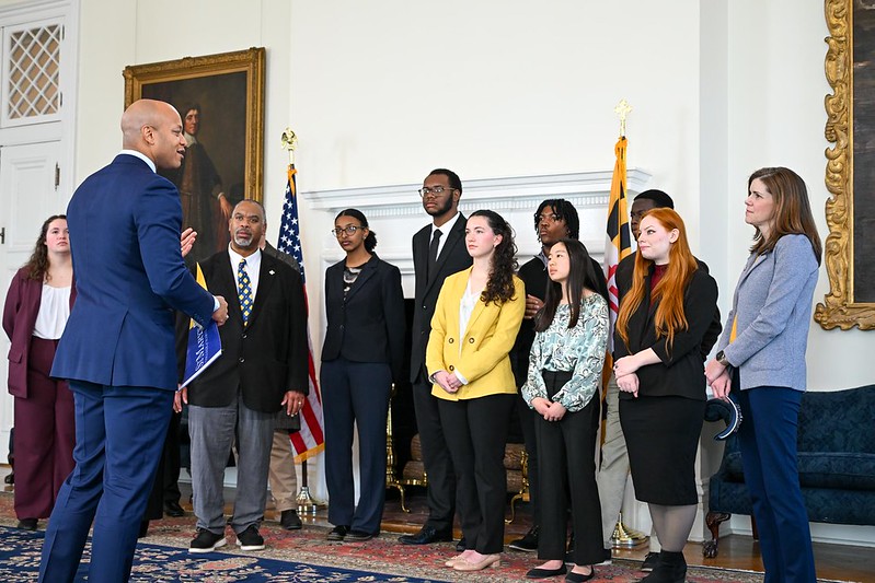 A man in a blue suit speaks to a group of eight people standing in a formal room with flags, a fireplace, and a portrait in the background.