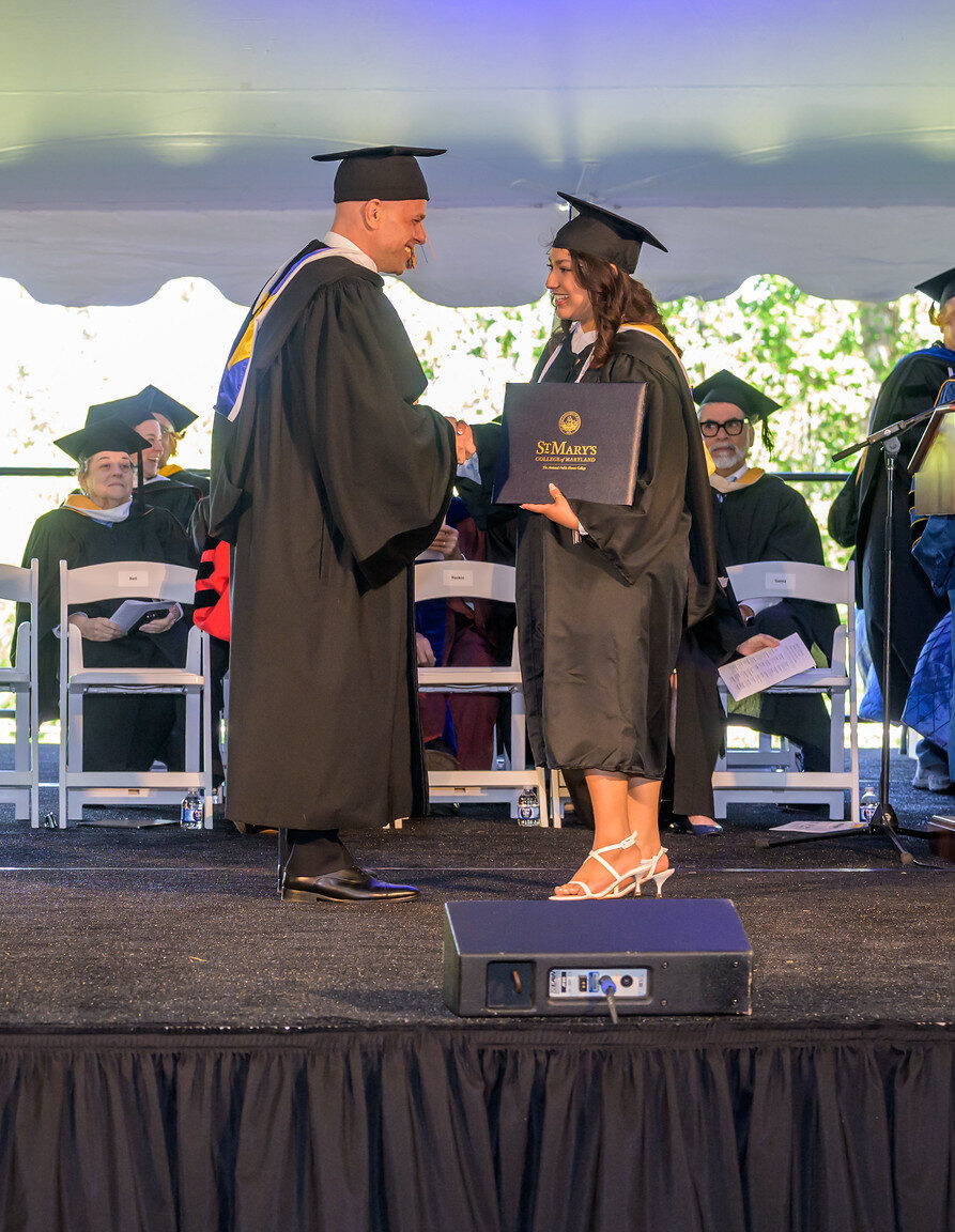 A graduate in cap and gown receives a diploma and shakes hands with a faculty member on stage during a graduation ceremony.