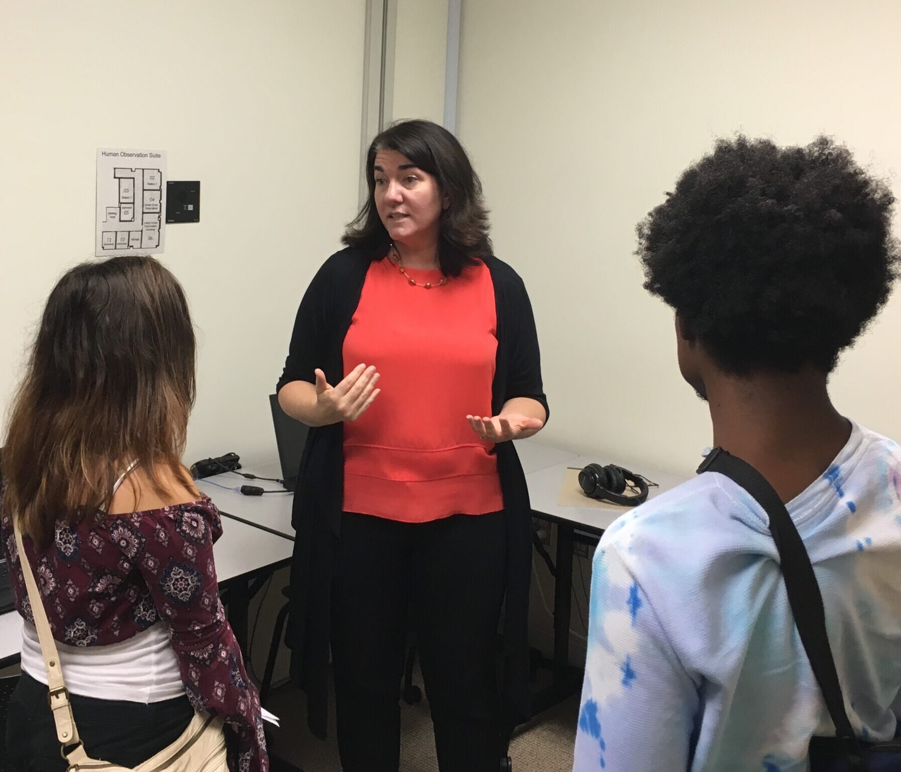 A woman stands in an office classroom, speaking to three students who are facing her. Computers and headphones are on desks in the background.