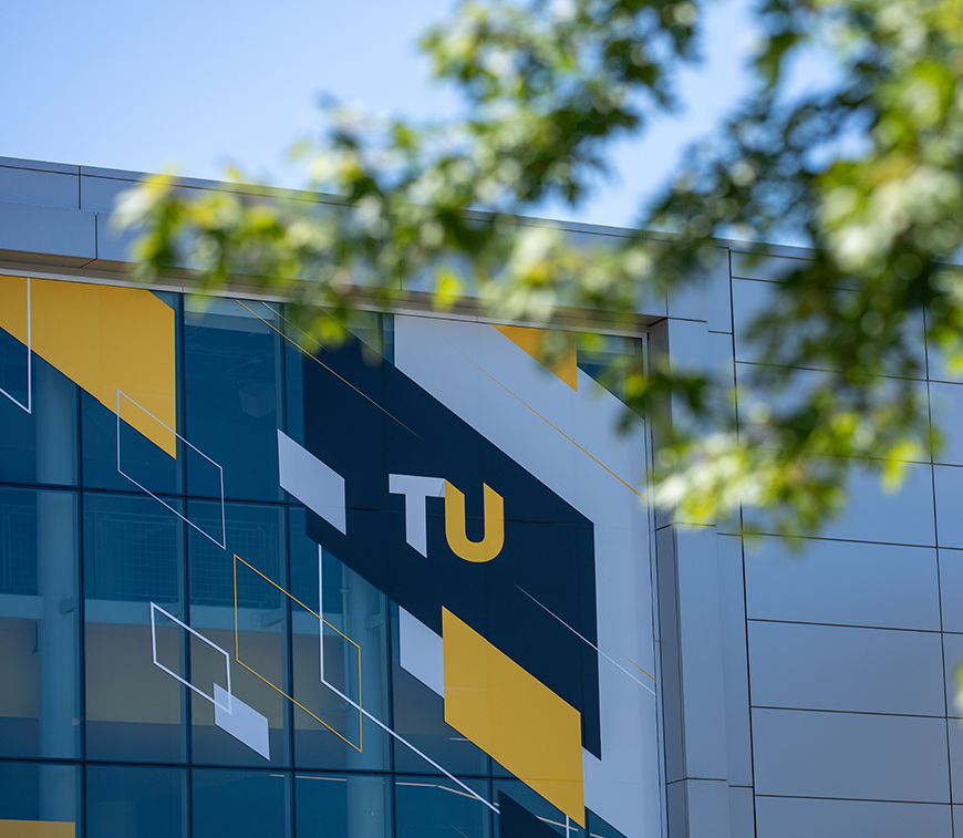 Modern building facade with large geometric mural in yellow, white, and black, featuring the letters “TU.” Tree branches partially obscure the view.