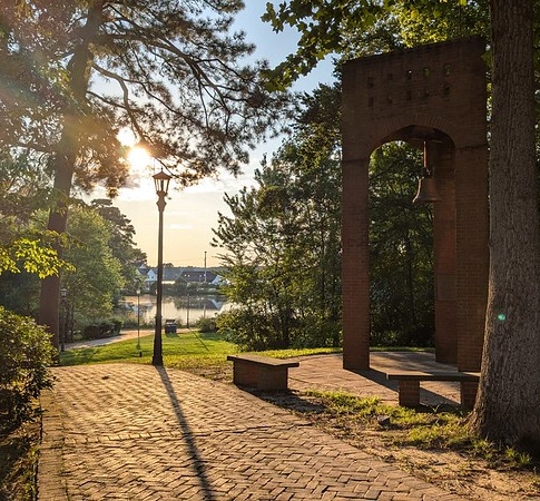 Brick arch with a bell stands in a shaded park area near a paved walkway, trees, benches, and a lamppost, with a river and sunlight in the background.